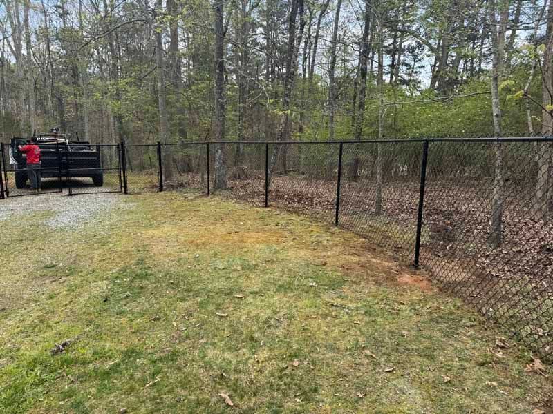 Chain-link fence along a grassy path beside woods, with two people near a black gate on the left.