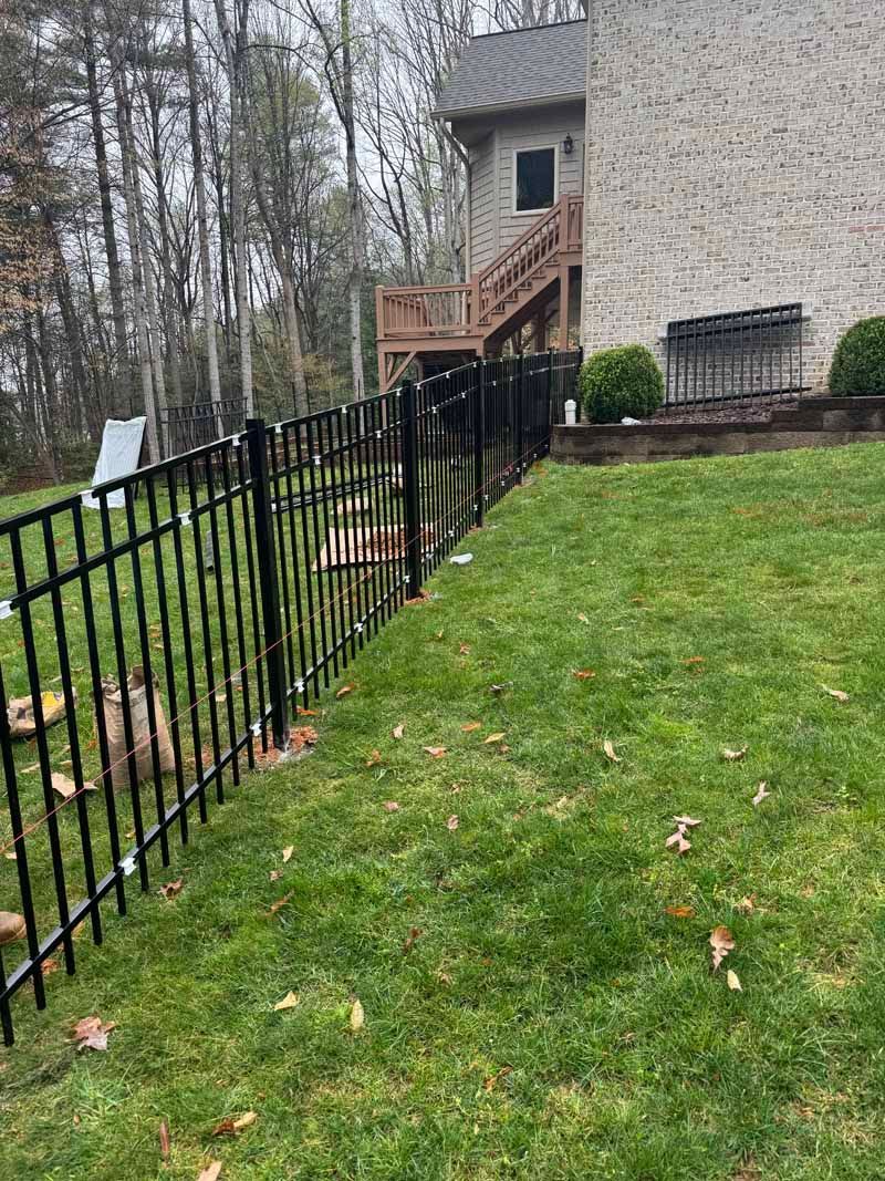 Black metal fence beside a grassy yard, leading to a brick house with wooden stairs and woods in back.