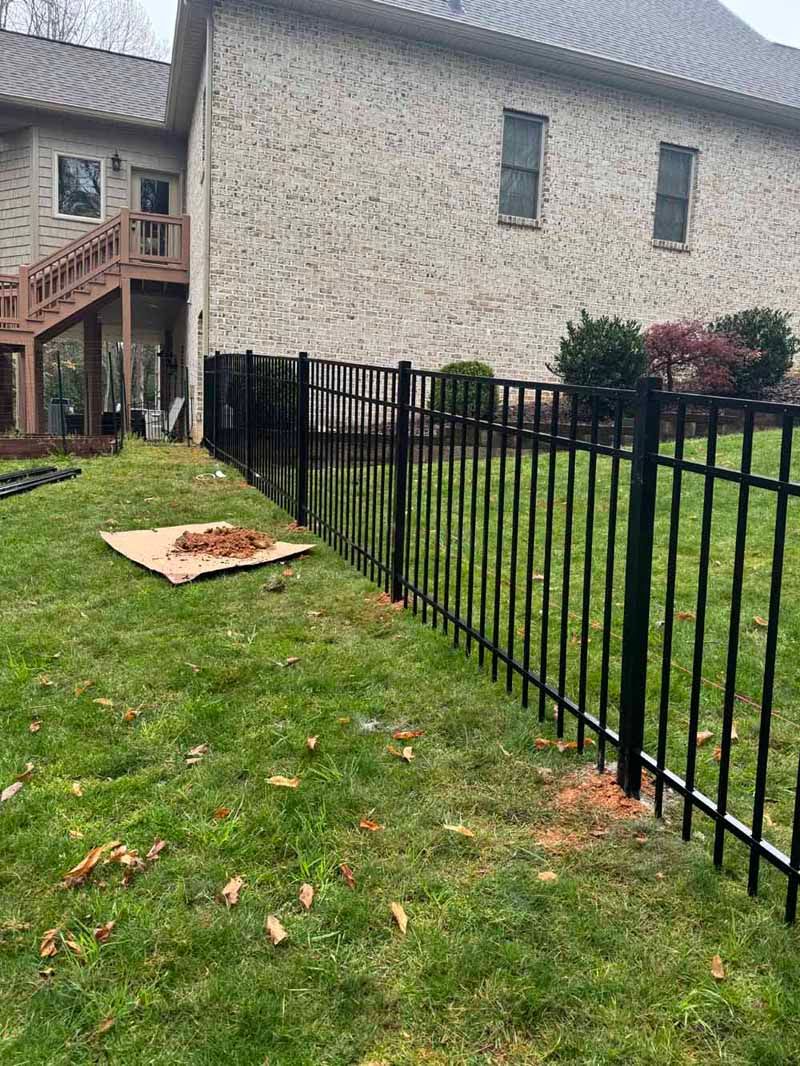 Black metal fence along a grassy backyard beside a beige house with stairs and a patio slab.