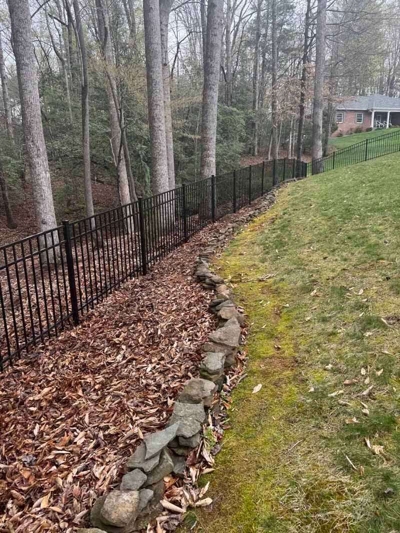 Backyard slope with black fence, leaf-covered ground, bare trees, and grassy lawn beside a house