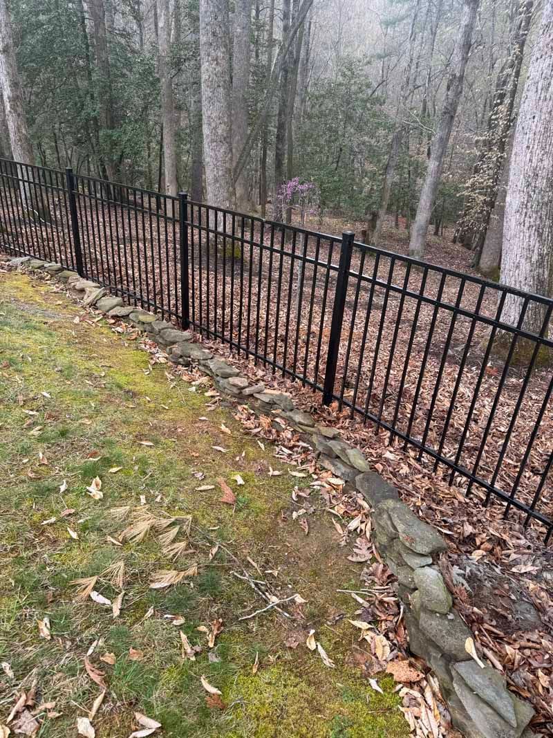 Black metal fence along a wooded slope with bare trees and fallen leaves