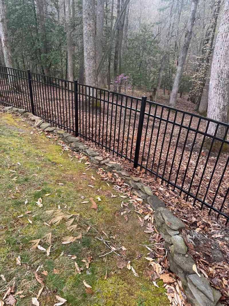 Black metal fence along a leaf-covered wooded slope beside a grassy path.