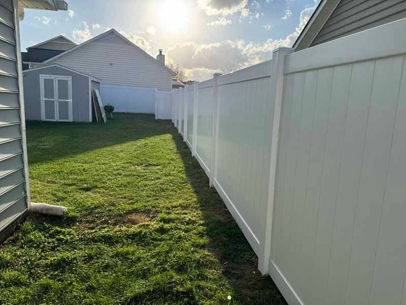 Narrow grassy backyard beside a white vinyl fence and house, sun shining from the far end.