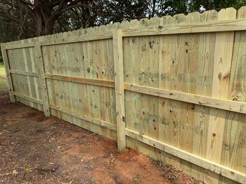 New wooden privacy fence along a dirt yard, with trees behind it.