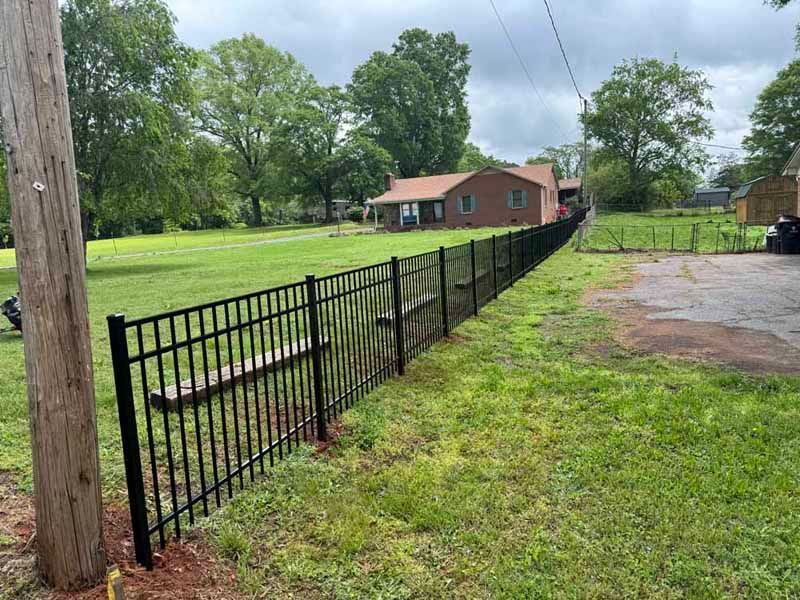 Black metal fence along a grassy yard beside a driveway, with a small house and trees in the background.