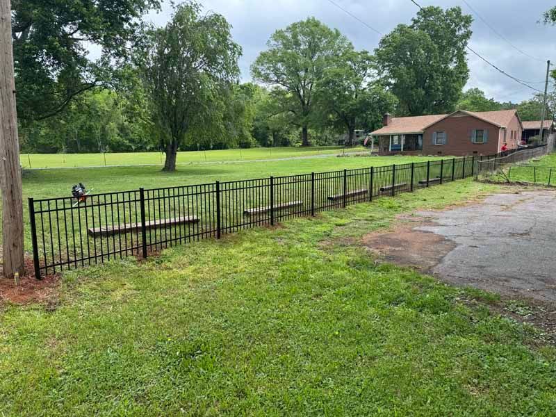 Black metal fence beside a grassy park with picnic tables, trees, and a small pink building near a paved road