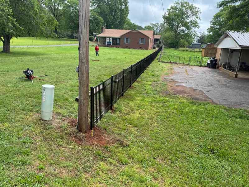 Backyard with black fence, red brick house, utility pole, and small shed under cloudy sky