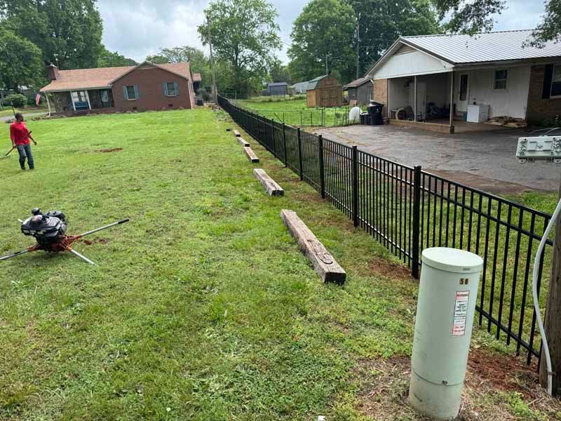 Backyard fence installation with workers, lawn tools, and a long black metal fence beside a house