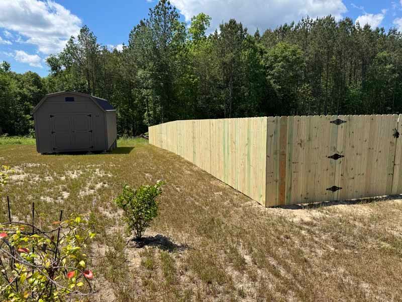 Backyard with wooden fence under construction, a dark shed, and trees under a blue sky