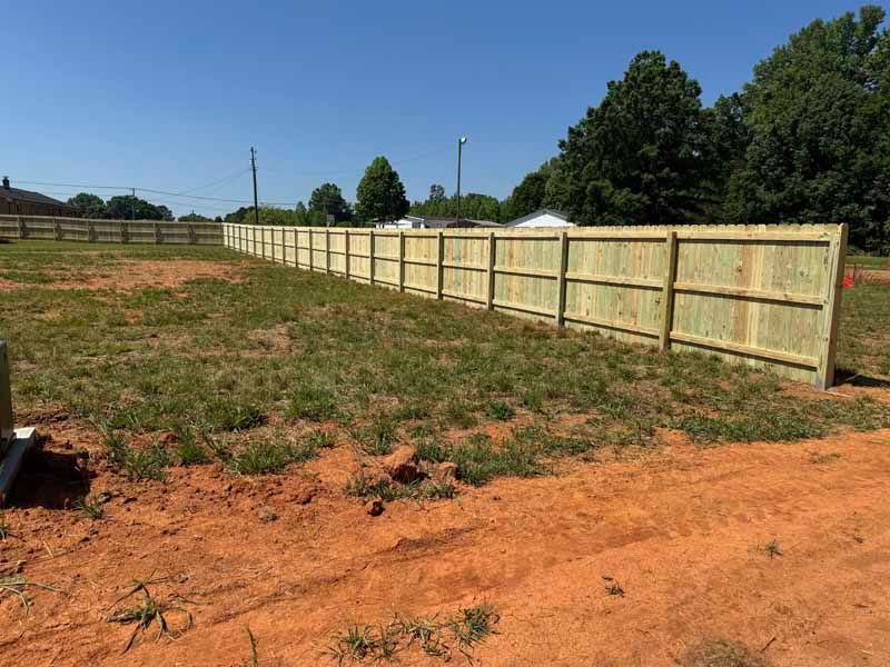 Backyard with a long wooden privacy fence, patchy grass, and red dirt under a clear blue sky