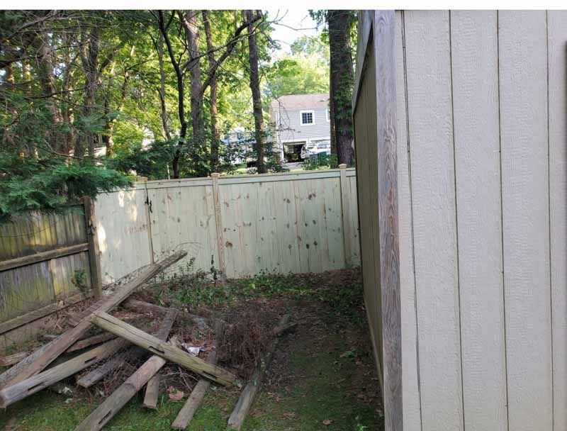 Backyard corner with scattered wooden boards, a fence, and a house in the background