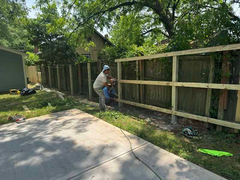 Person installing a wooden fence beside a driveway in a leafy yard