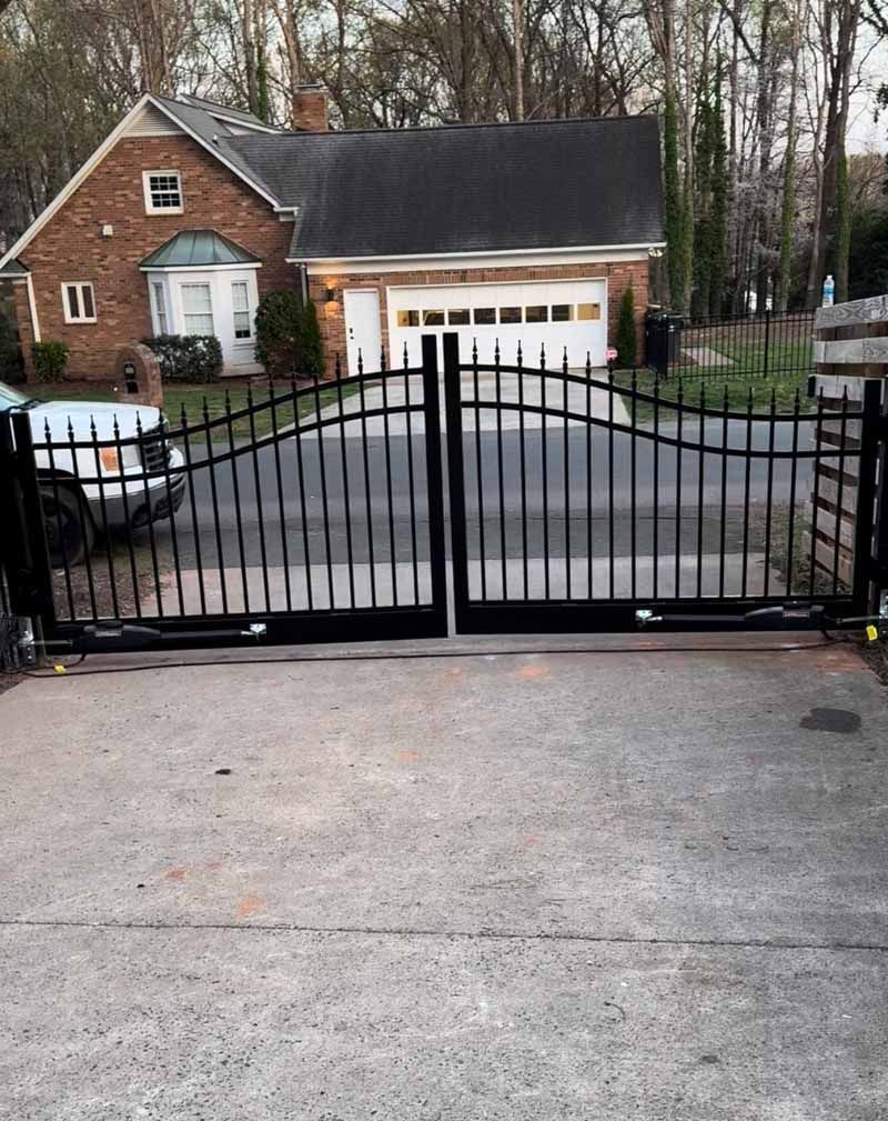 Black metal driveway gate in front of a brick house with a garage and trees.