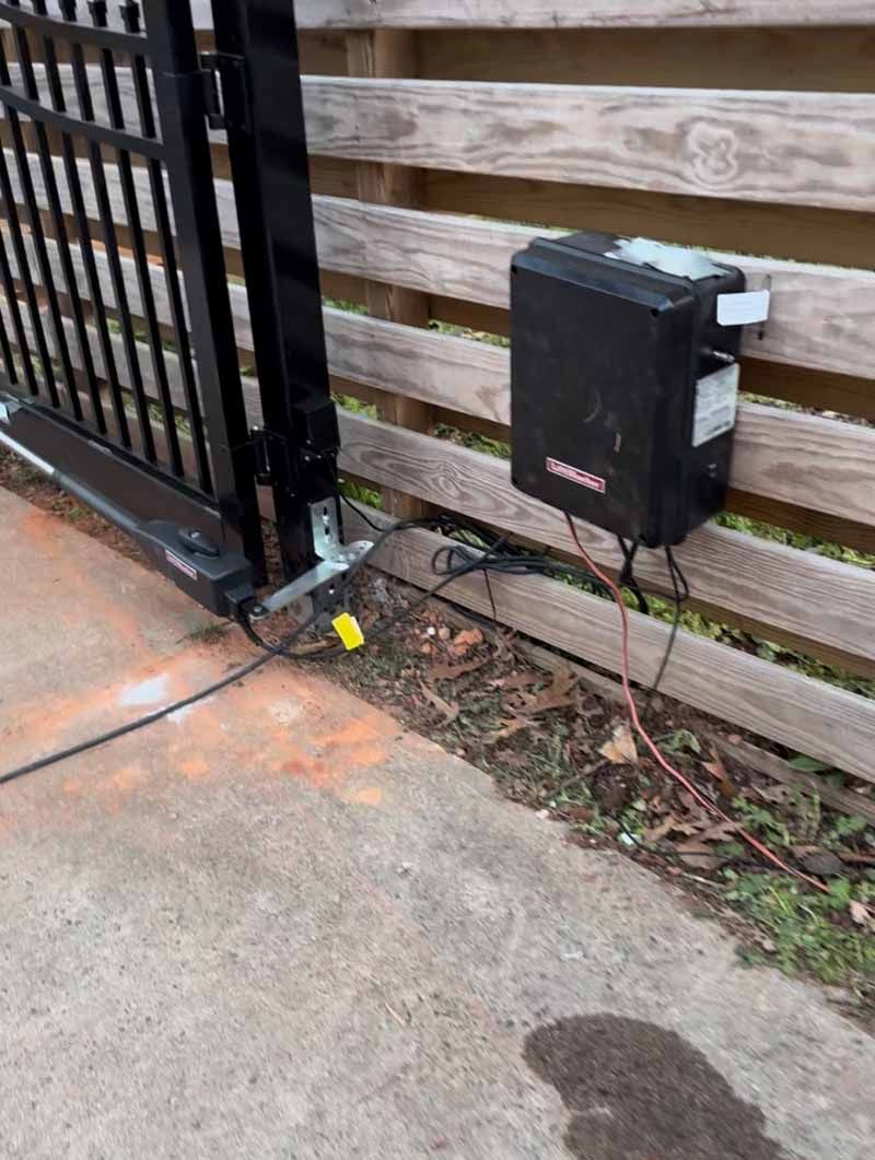 Black gate motor and control box beside a wooden fence on a concrete driveway.