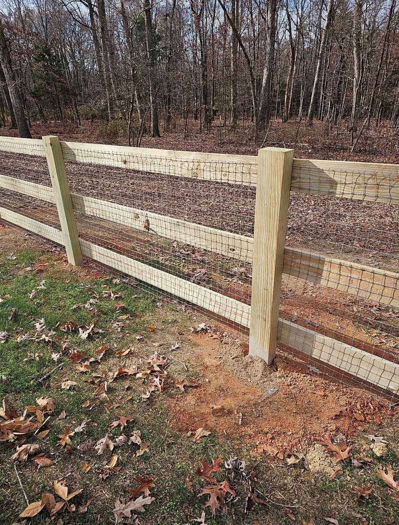 Wooden split-rail fence along a wooded path with dry leaves on the ground.
