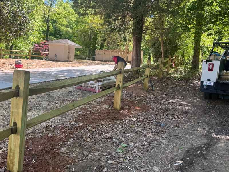 Person working by a wooden fence near a wooded driveway, with a truck and small building in the background.