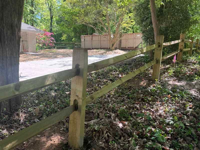 Wooden fence beside a gravel driveway in a leafy yard with a tan gate and trees.