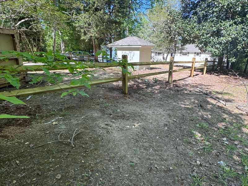 Wooden fence along a shaded dirt path with a small white shed in a wooded yard.