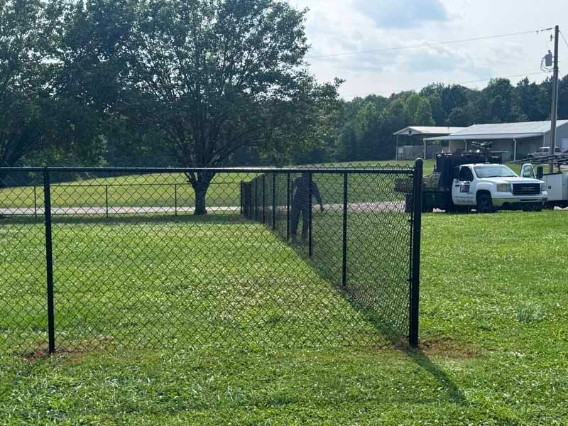 Chain-link gate in a grassy yard, with a tree and white truck near a building in the background.