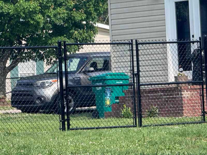 Parked dark SUV beside a green trash bin behind a chain-link fence in a suburban yard