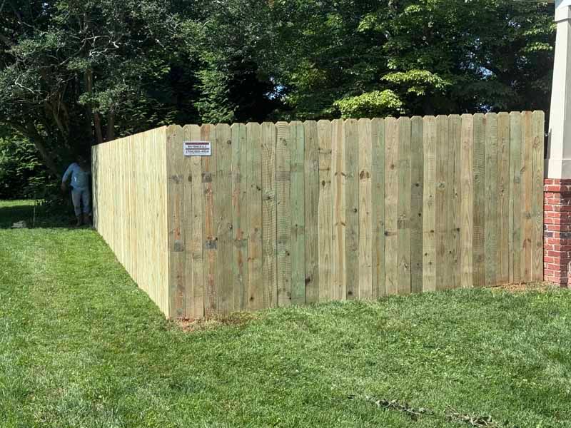 Wooden privacy fence enclosing a yard beside a grassy lawn and trees, with a brick wall at right.