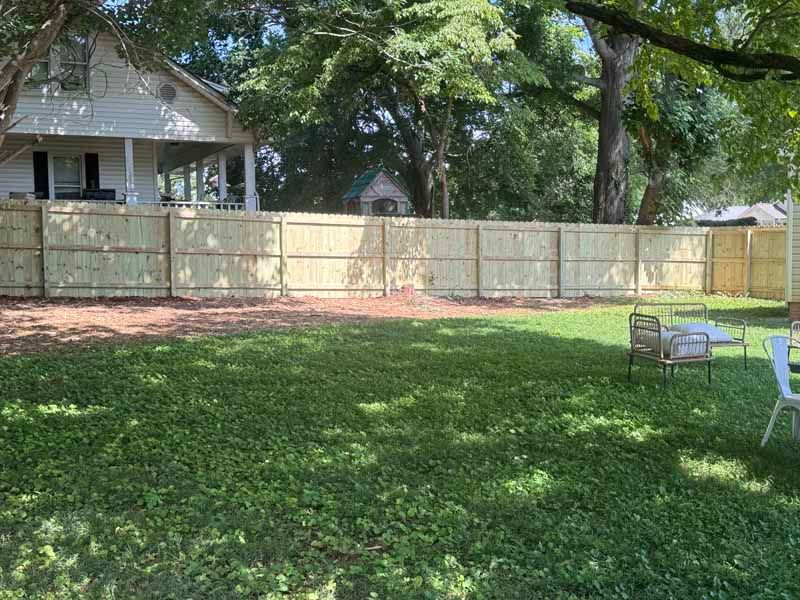 Backyard with a wooden fence, large trees, and lawn chairs on green grass