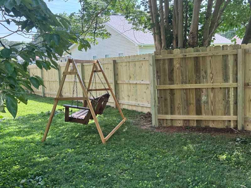 Wooden swing bench under trees in a grassy backyard beside a tan privacy fence.