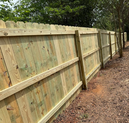 Wooden privacy fence along a wooded dirt path, with trees in the background.