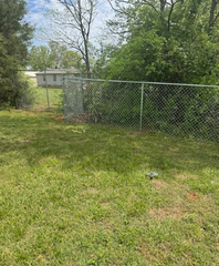 Chain-link fence along a grassy yard with trees and a small building in the background.