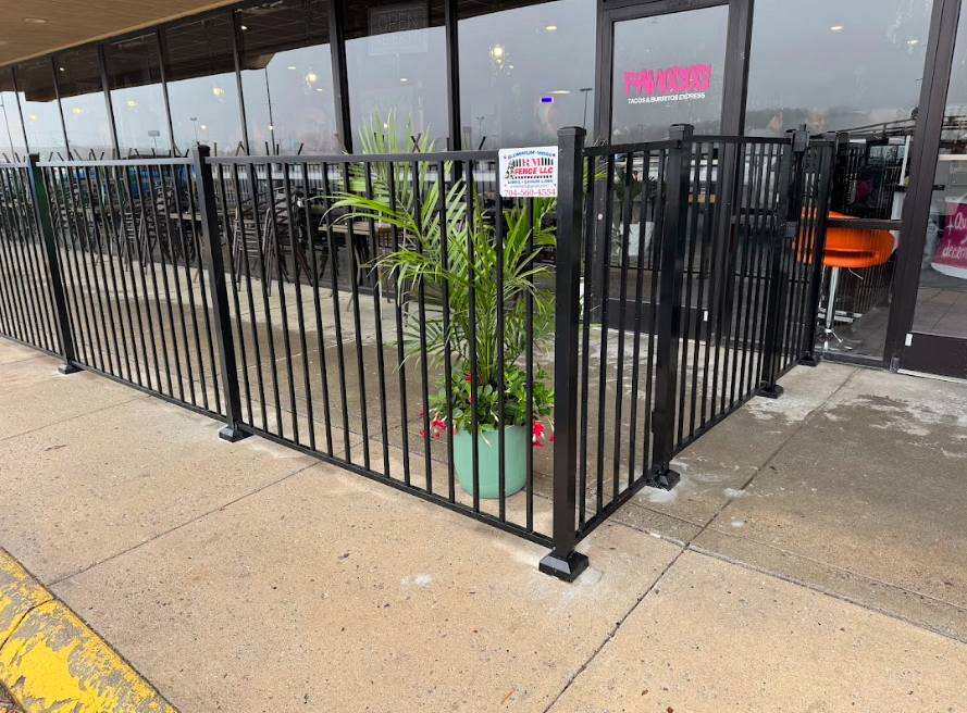 Black metal fence enclosing a restaurant patio with tables, chairs, and potted plants by the sidewalk
