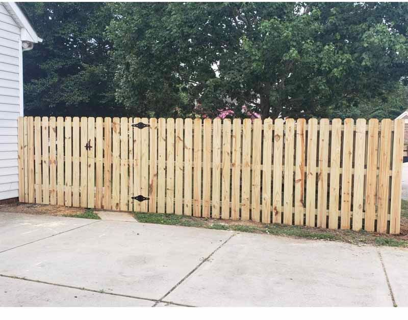 Light wood fence along a concrete driveway, with trees and a house beside it.
