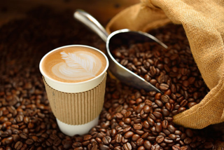 Latte in a to-go cup, sitting amid roasted coffee beans, with a scoop and burlap sack in the background.