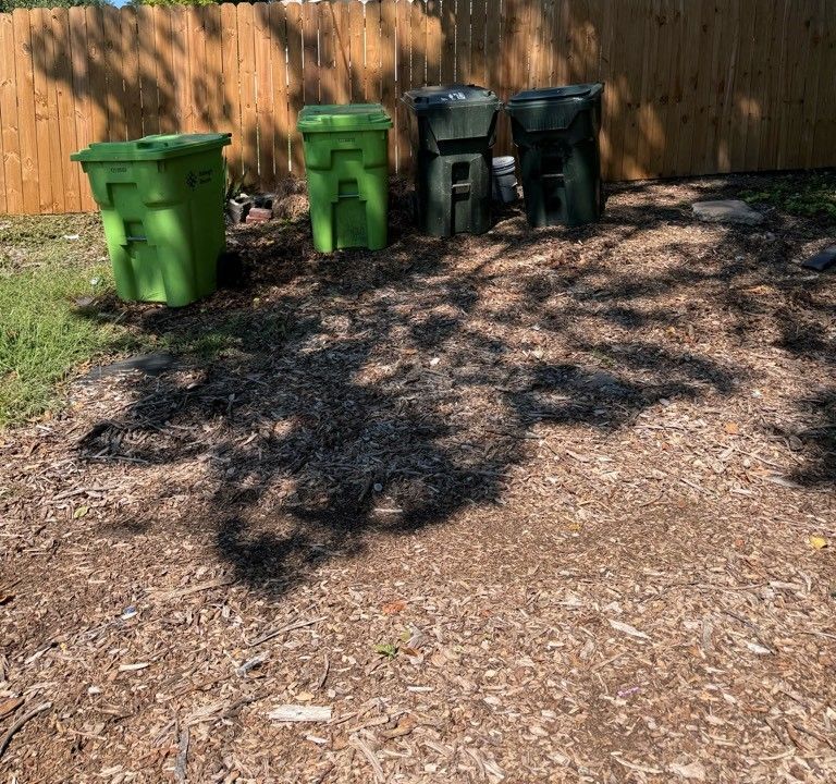 Three green trash cans are sitting on the ground in front of a wooden fence