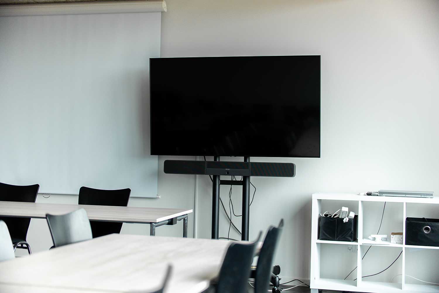 Mødelokale med stor tv-skærm, soundbar, borde, stole og opbevaringsrum.
