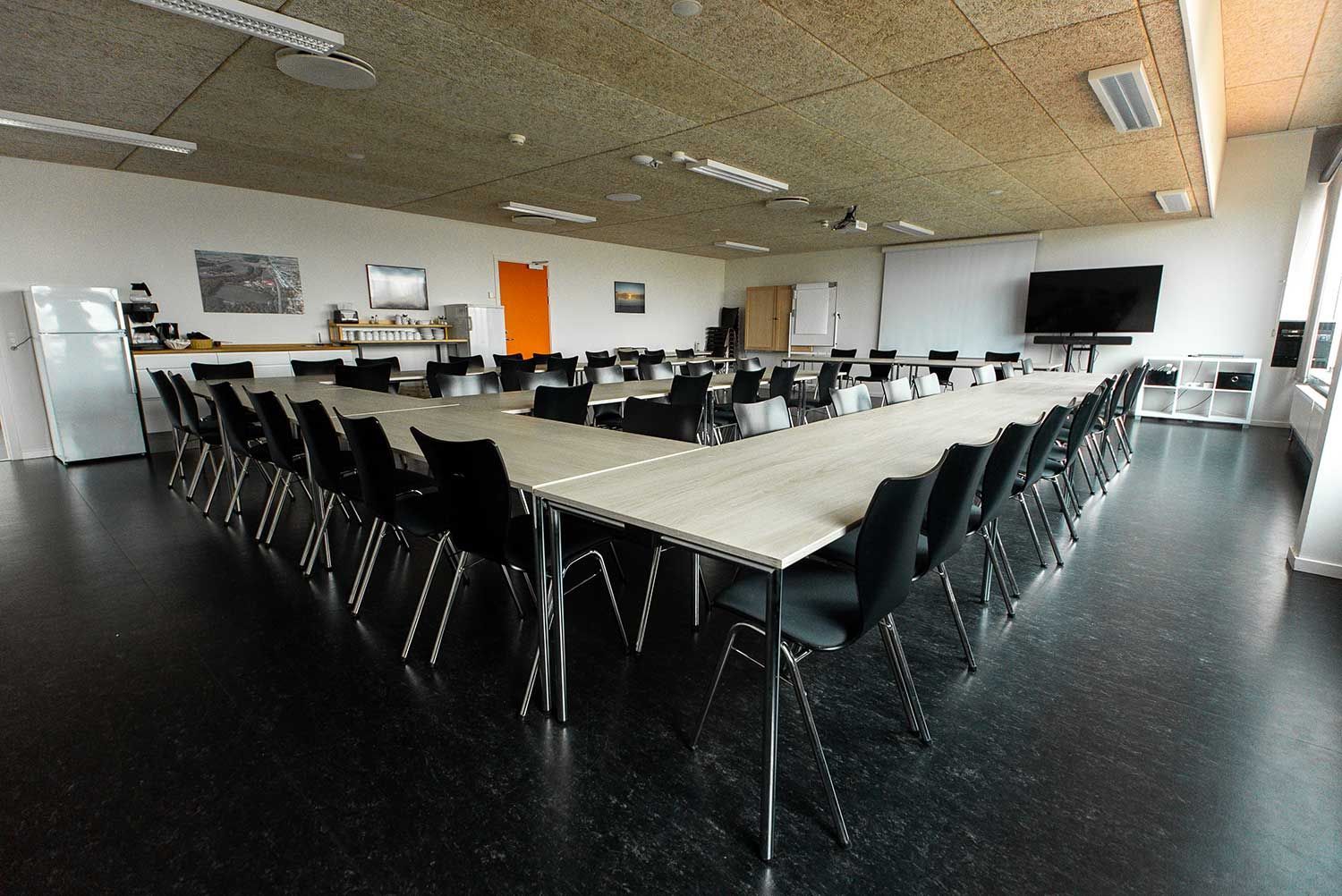 Konferencerum med U-formet bord, sorte stole, mørkt gulv og TV på væggen.