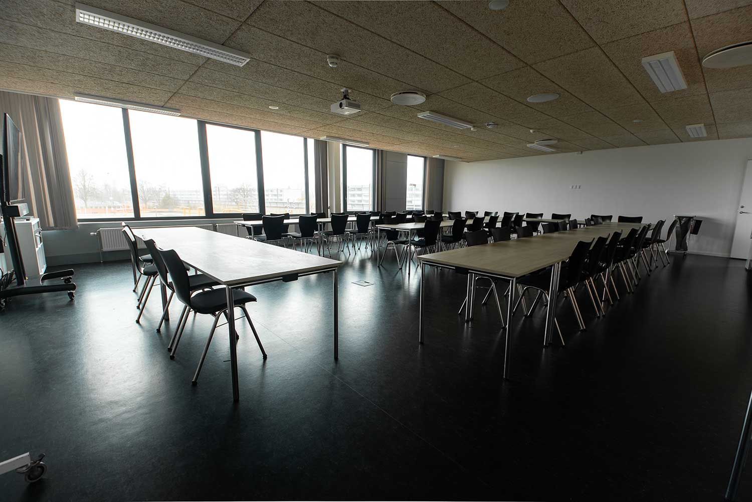 Tomt konferencerum med borde og stole arrangeret i en U-form, store vinduer og mørke gulve.