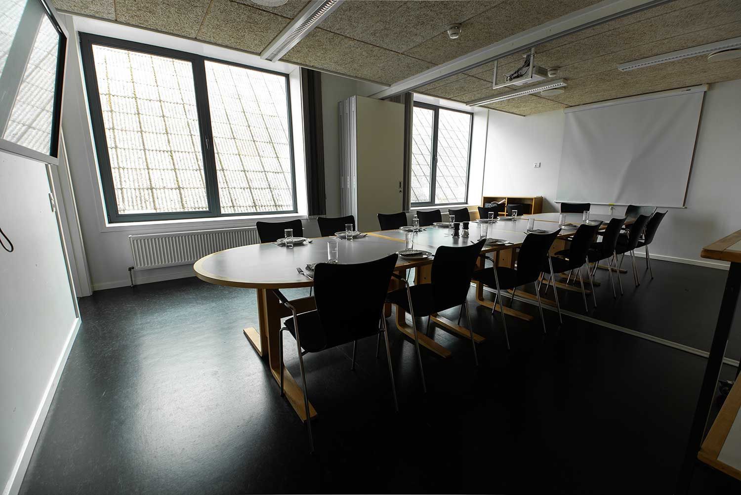Konferencerum med ovalt bord og sorte stole, med vinduer og projektorlærred.