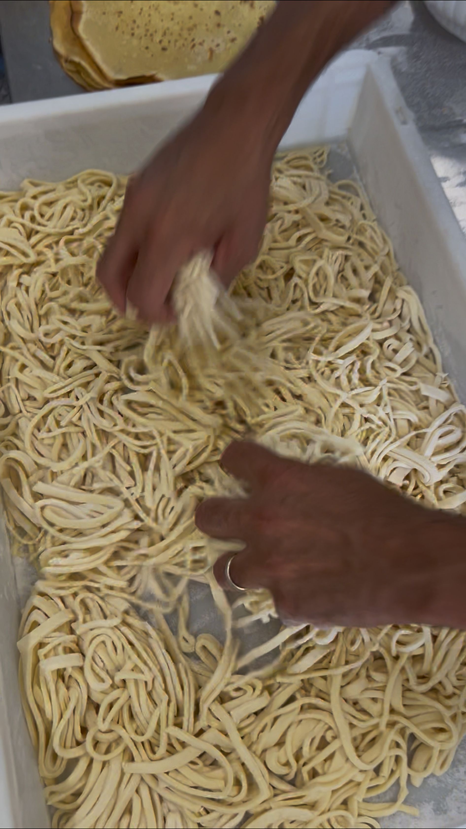 Mani che impastano fili di pasta in un grande vassoio, preparando noodles o pasta fresca