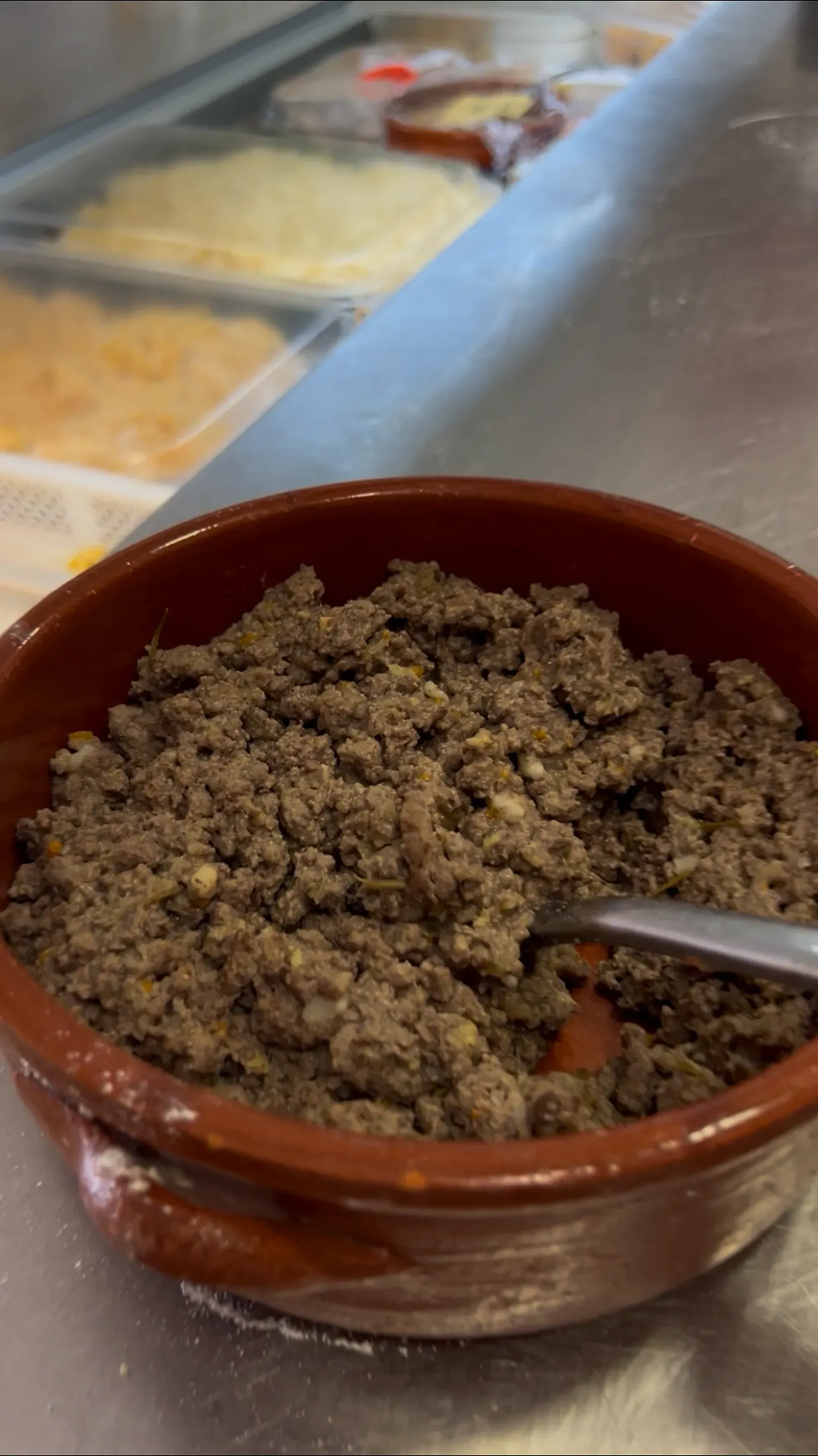 Ciotola marrone con carne sbriciolata e un cucchiaio su un bancone da buffet, con vassoi di cibo sullo sfondo