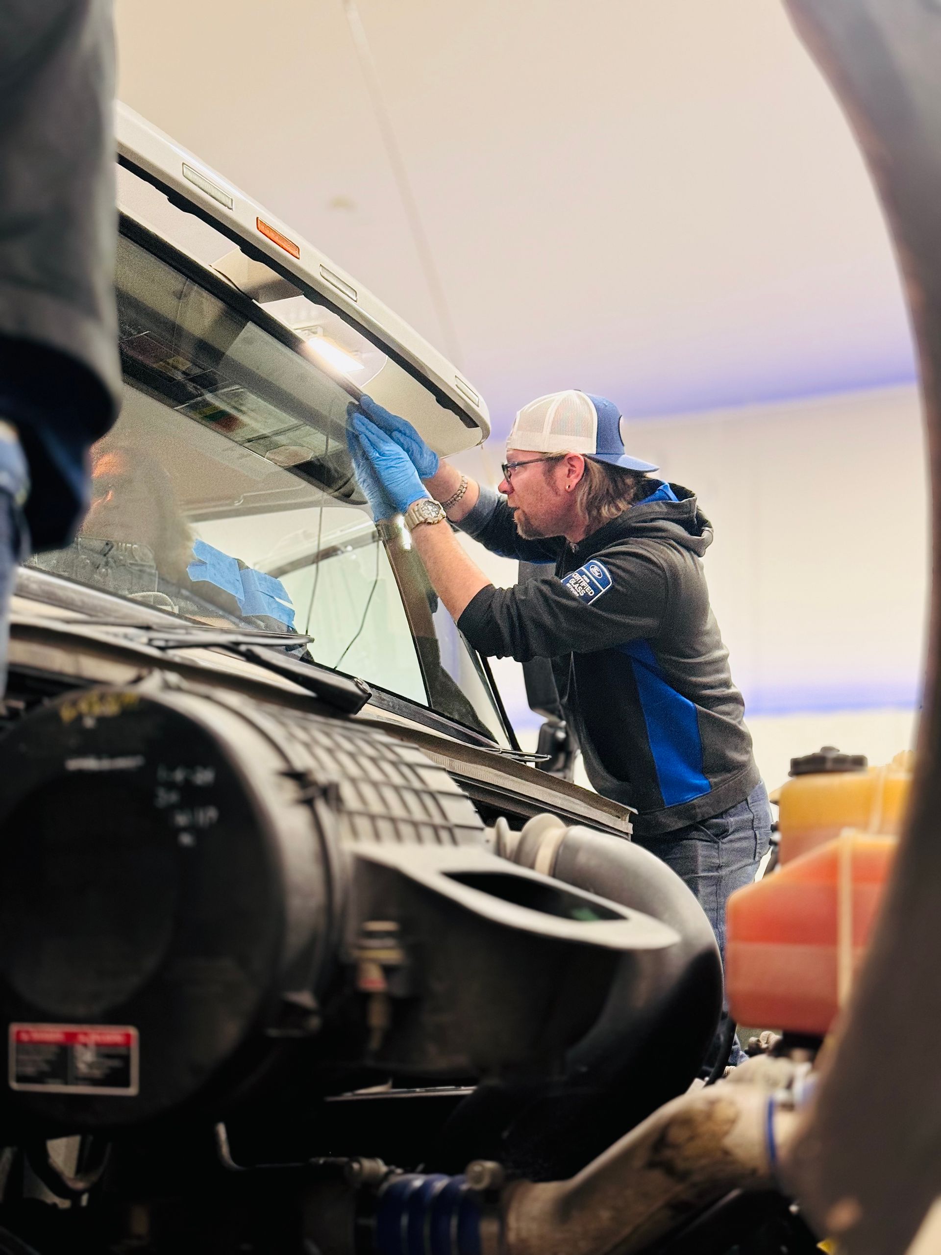 Person in a blue-accented hoodie cleaning a truck's windshield in a garage, wearing gloves and a baseball cap.