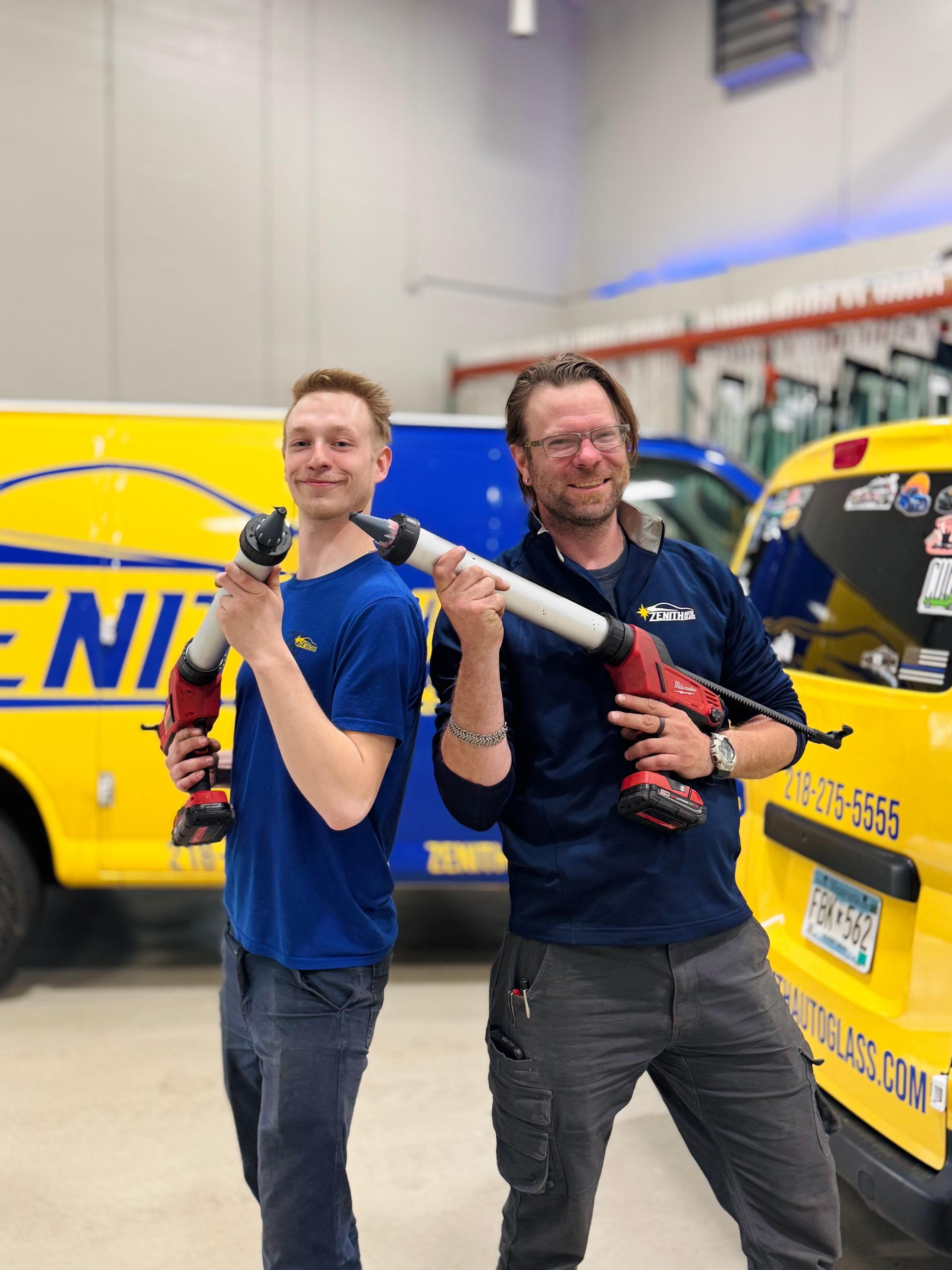 Two men holding caulking guns near yellow work vehicles.