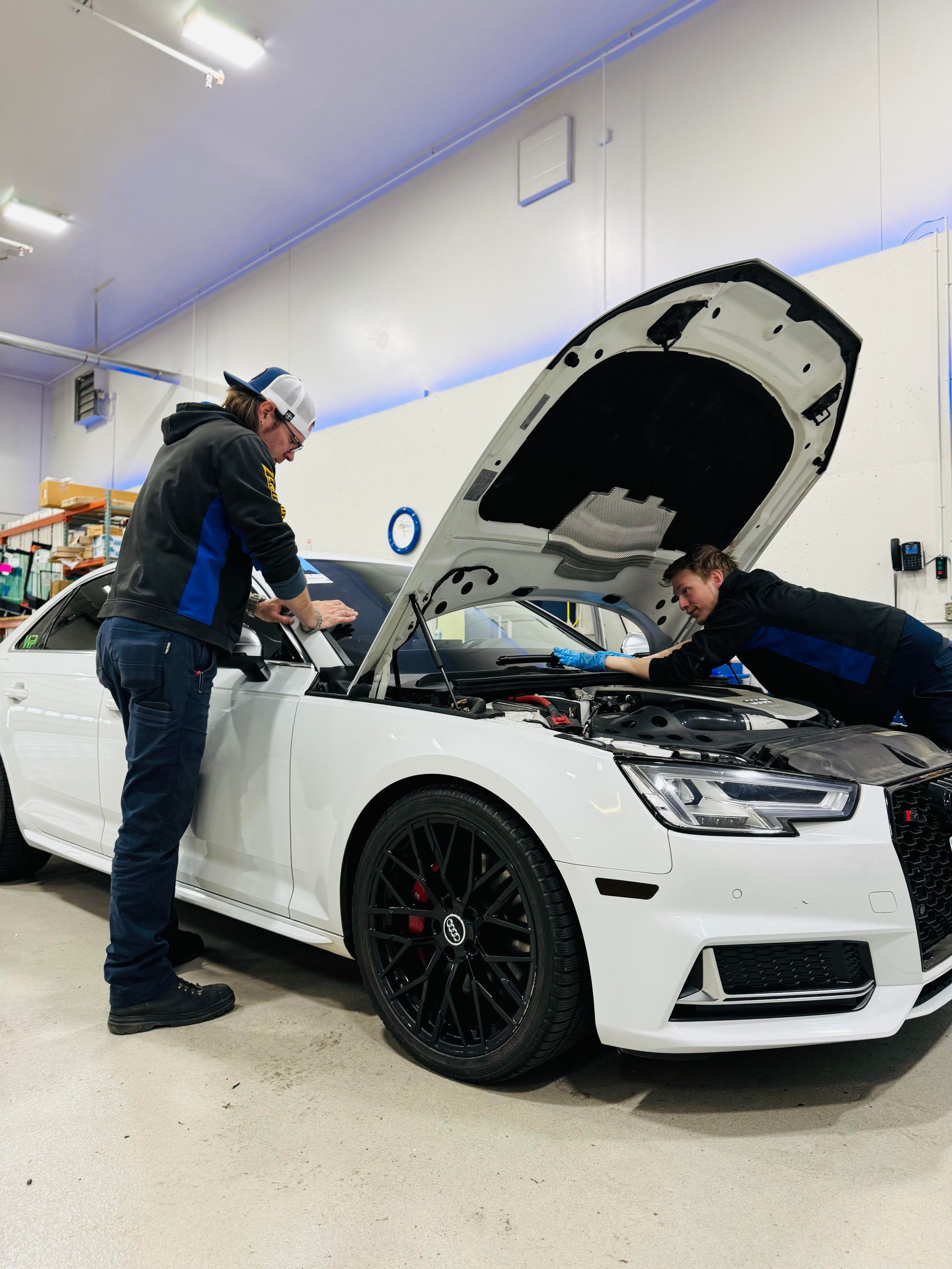 Two mechanics working on a white car with an open hood in a garage.