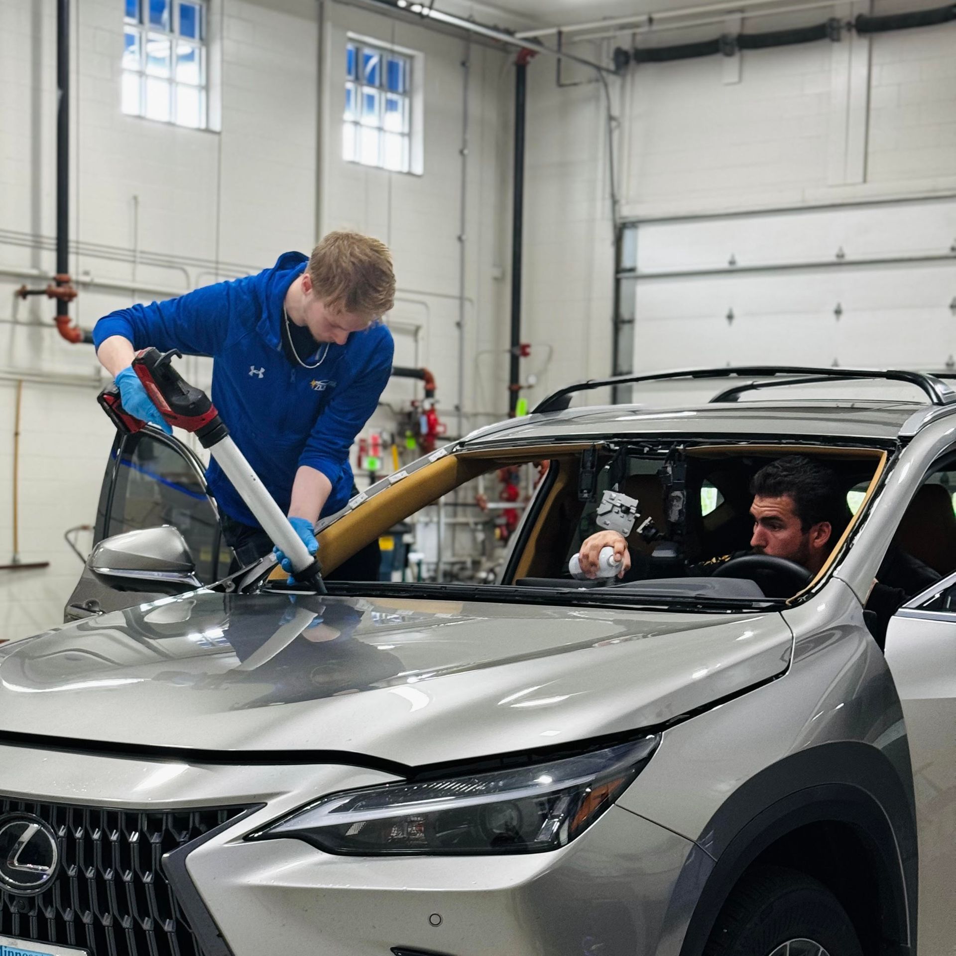Two people replacing a windshield on a silver Lexus in a repair shop. One applies sealant, the other inside the car.
