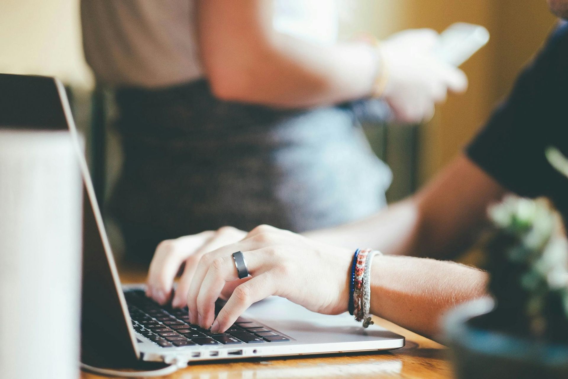Person typing on laptop, other person in background holding phone.