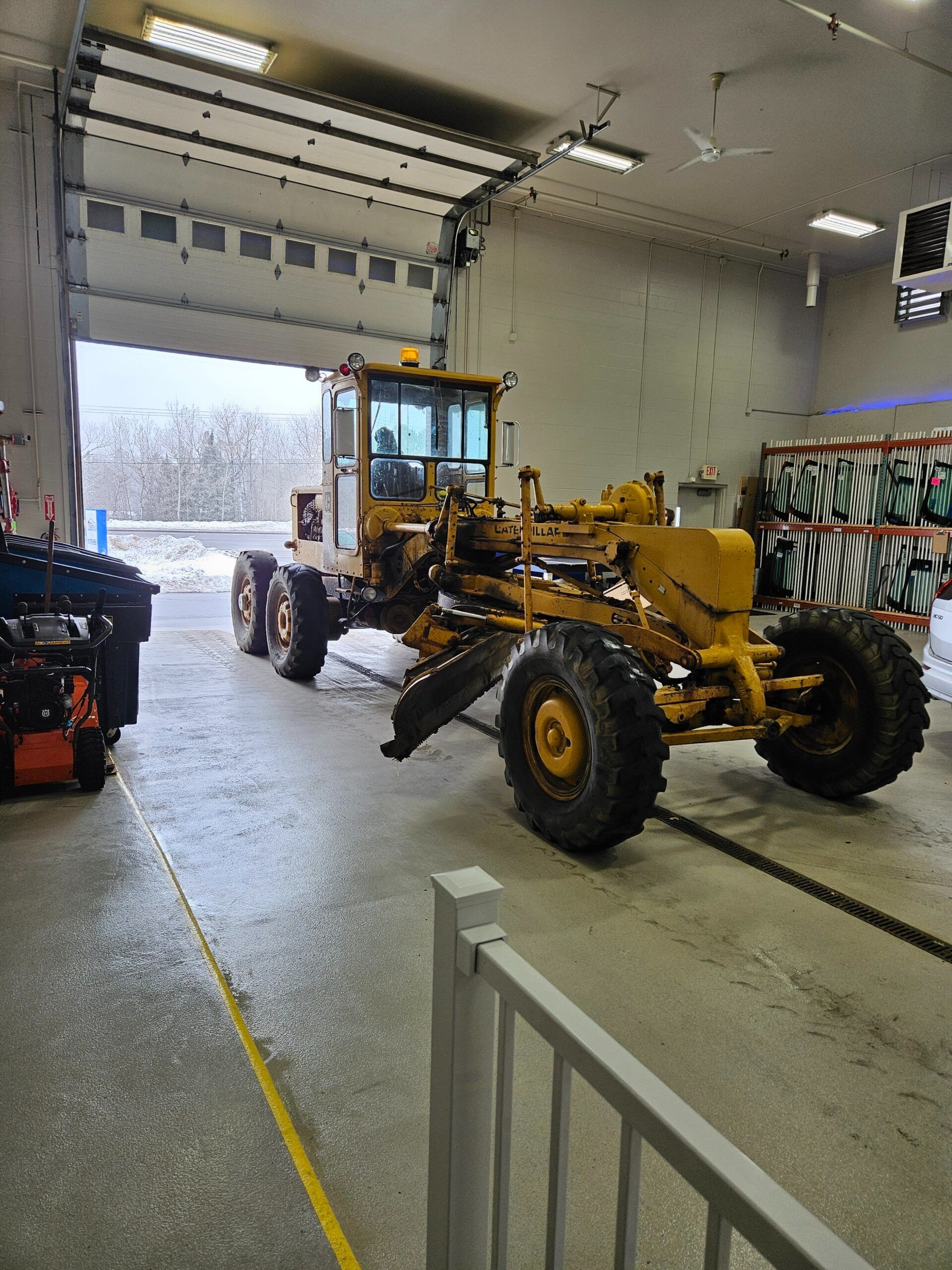Yellow motor grader inside a garage with an open door; snow is visible outside.