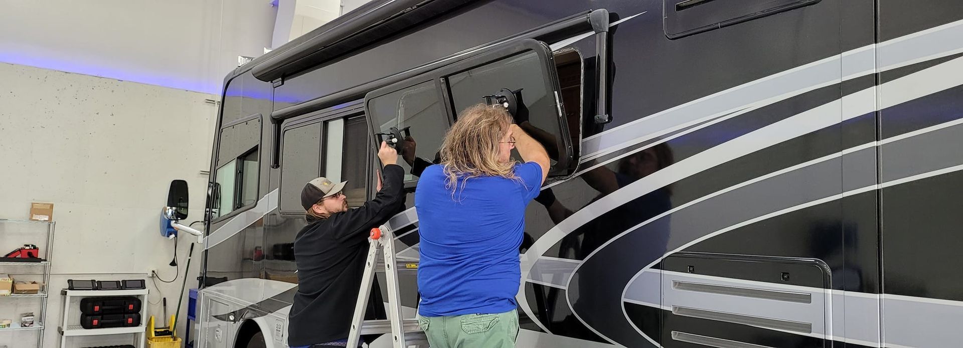 Two people working on the side of a black and silver RV, one on a ladder.