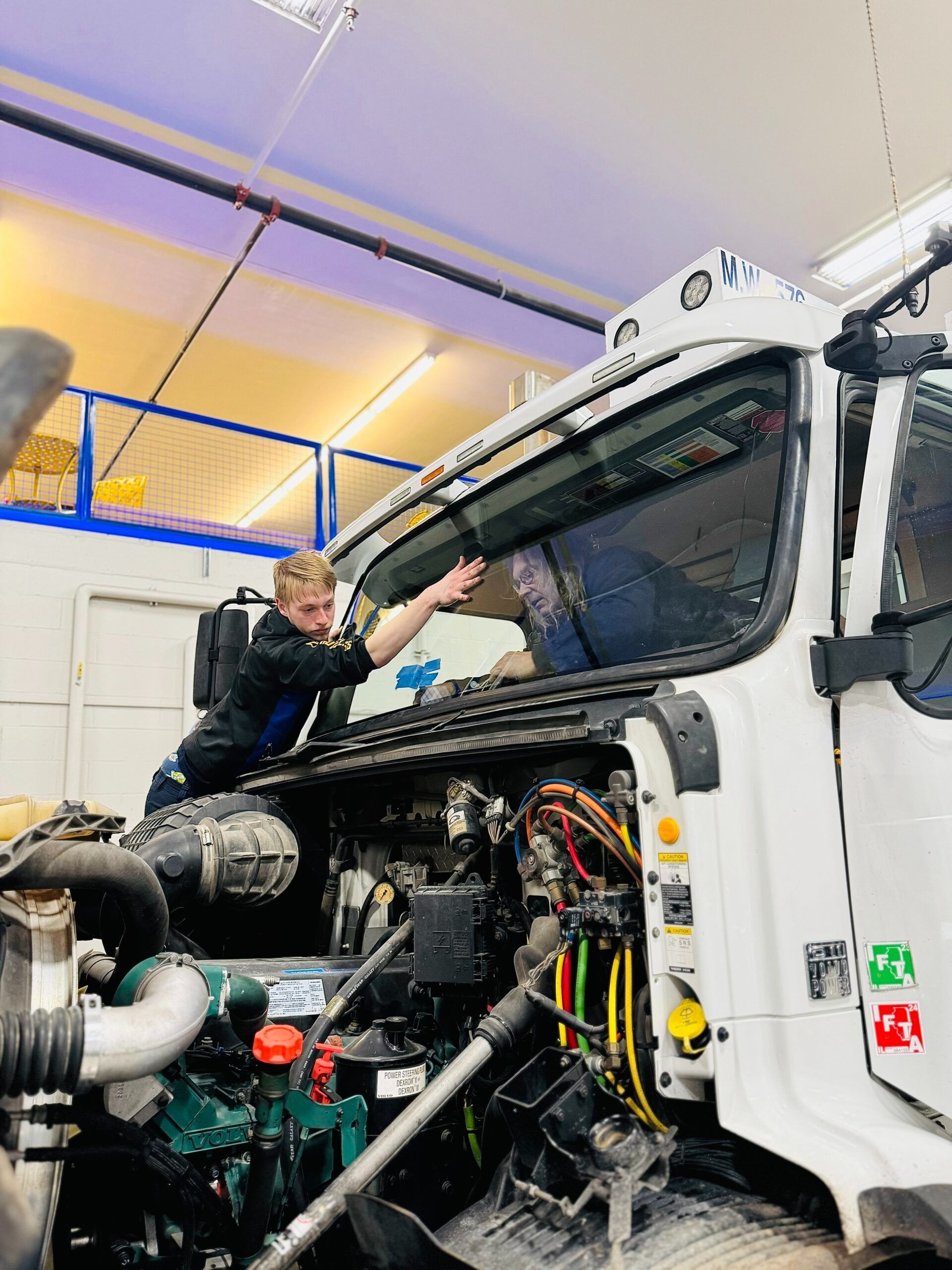 Mechanic working on the windshield of a white semi-truck in a garage, reaching with hands.