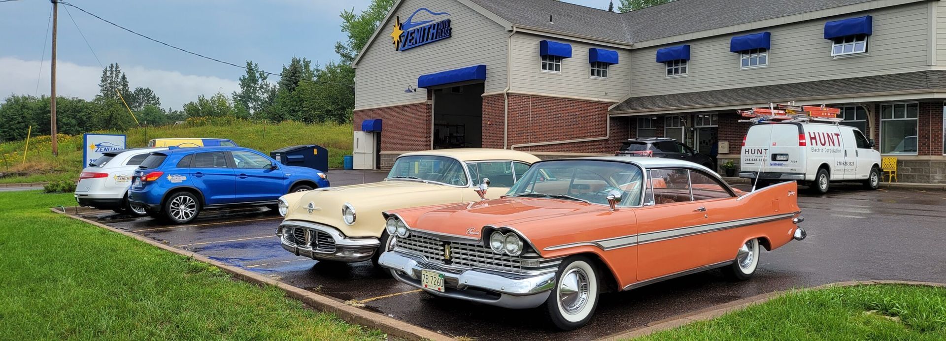 Classic cars parked outside a building. One is orange, and another is light yellow. Several other cars sit in the lot.