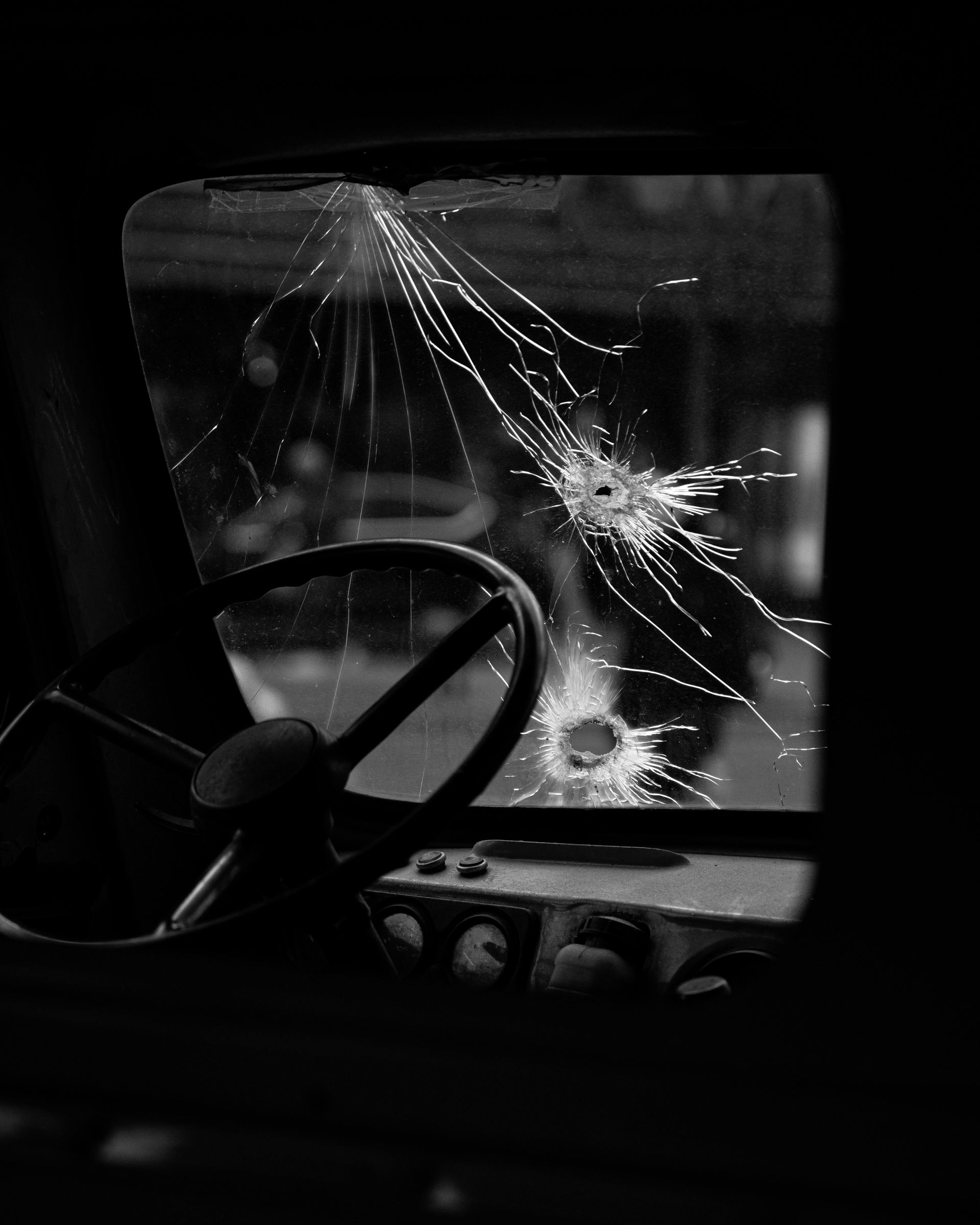Black and white interior shot of a vehicle, steering wheel visible. Windshield is shattered.