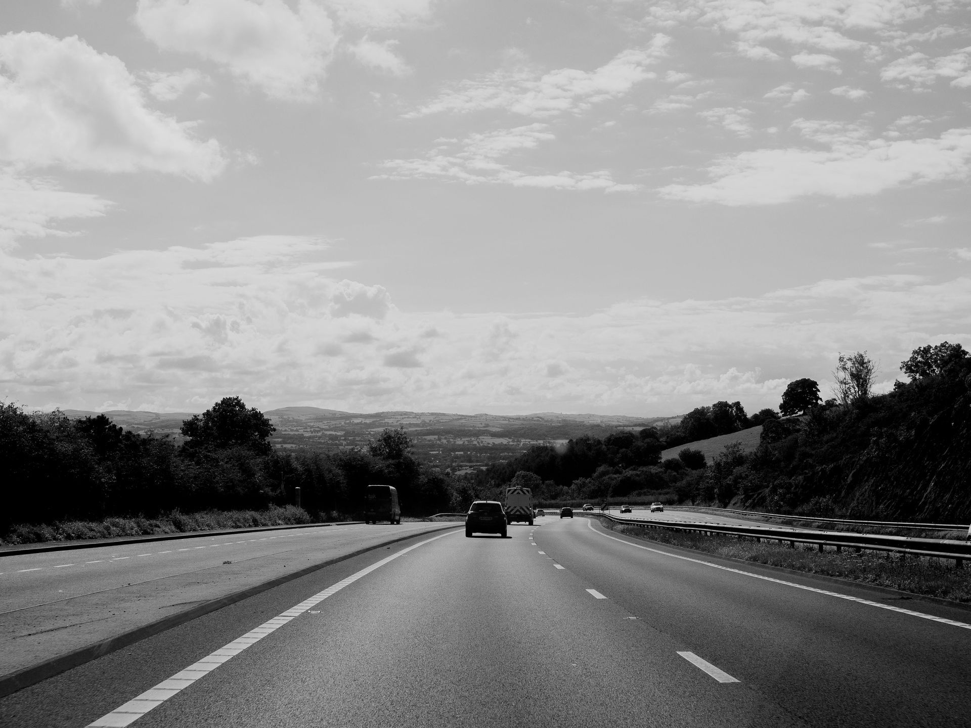Black and white image of a highway with cars driving toward a distant town under a cloudy sky.